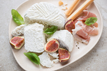 Ricotta cheese with figs, prosciutto, grissini and green basil on a beige plate, horizontal shot, middle close-up, selective focus