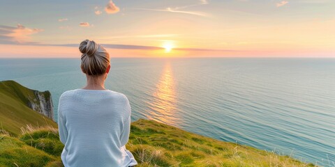 Woman sitting on a grassy cliff, watching a beautiful sunset over the ocean horizon, signifying peace and tranquility.