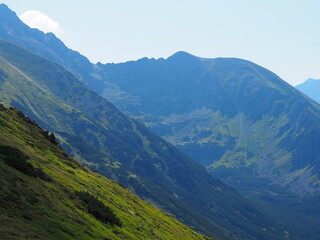 Obraz premium An expansive view of a mountain range with rocky peaks and lush greenery under a clear sky, showcasing the grandeur and unspoiled beauty of the natural environment.