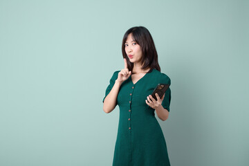 A woman in green dress playfully holds phone and gestures for silence, conveying sense of secrecy and intrigue.