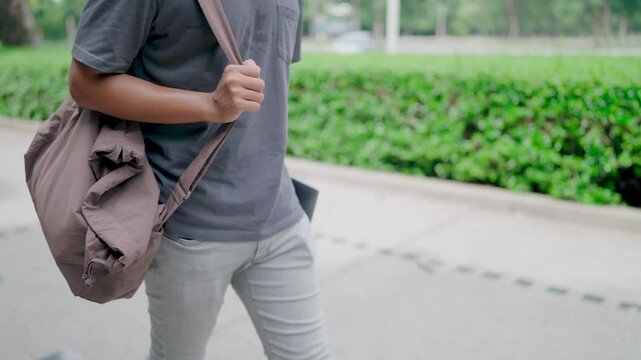  Asian man in gray jeans walking with laptop and bag in hand in university park. Walking to study. He is walking in nature to relax from work and study. Study online. Freelance work.