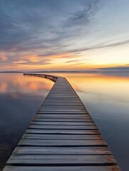 Fototapeta premium A wooden pier extends into a calm lake at sunset, creating a path towards a horizon of vibrant colors and clouds.
