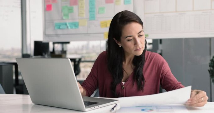 Office employee using laptop, checking, cross-referencing information from papers filled with charts and diagrams, analyzing market trends, updating report, preparing presentation for upcoming seminar