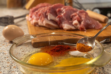 Cook adds ground red pepper to a plate with broken raw eggs to make a liaison, with beaten pork tenderloin and a meat mallet in the background. Close-up