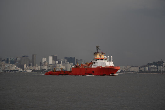 A bright red offshore supply vessel (OSV) navigates the calm waters near a city skyline, against a dark cloudy sky before storm. The ship's details, cranes, and deck equipment are clearly visible
