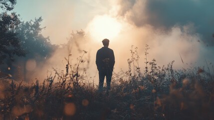 A silhouette stands amidst tall grass and wild foliage, facing the rising sun, enveloped by a soft mist and the tranquility of early morning