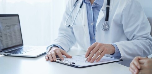 A doctor and a patient. The physician, wearing a white medical coat over a blue shirt, is filling...