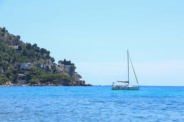 Sailboat floats on calm blue sea near rocky coastline, with hillside covered in greenery to the left under a clear sky. The composition highlights peaceful ocean and nature.