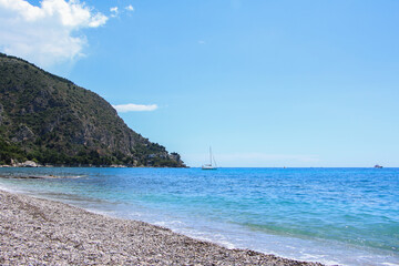 Pebble beach in Èze, French Riviera with turquoise Mediterranean Sea, sailboat on horizon, and forested cliffs. Ideal for travel, coastal tourism, and nature-themed visuals.