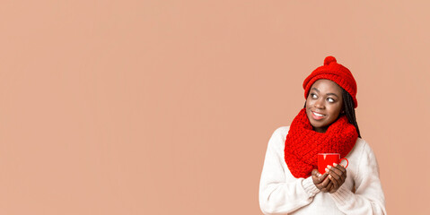 Interested african american winter woman enjoying hot tea or coffee, holding red mug, looking at copy space, beige background, panorama