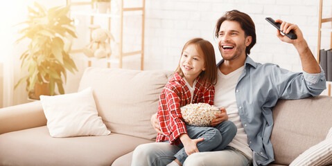 Little girl and her happy father watching football match on TV, celebrating success of their team, sitting on sofa with popcorn, family traditions concept, copy space