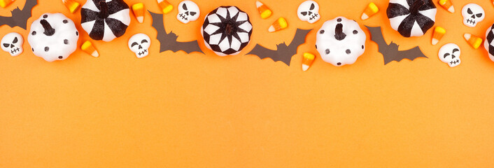 Halloween top border of black, white and orange color candy, bats and pumpkin decor. Above view on an orange banner background with copy space.