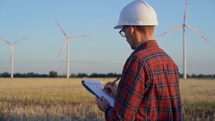 Man engineer, wearing a white protective helmet is taking notes with a clipboard in a field with wind turbines, as the sun sets. Clean energy and engineering concept