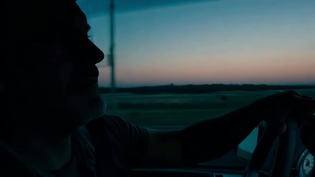 A man is seen driving through a serene landscape as the sun sets, deep in thought and enjoying the tranquility of the road. The soft hues of twilight create a reflective atmosphere.