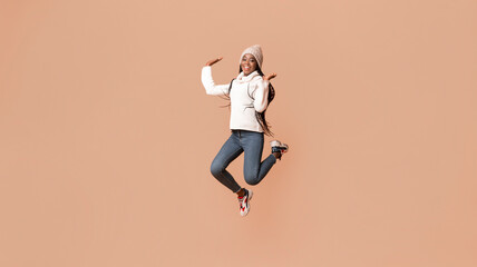 Fototapeta premium Playful young african woman in winter hat having fun over beige studio background, jumping in the air gracefully