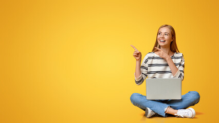 Cheerful Teenage Girl Sitting With Laptop On Her Laps And Pointing At Copy Space On Yellow...