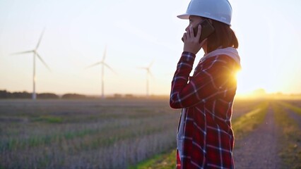 Female engineer talking on the phone in a field with wind turbines, as the sun sets. Concept of clean energy and engineering audit