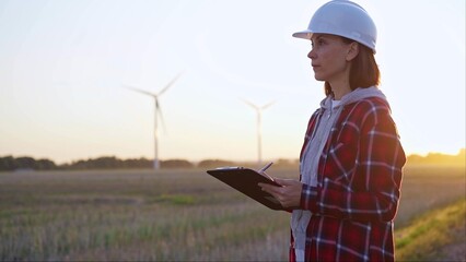 Adult woman engineer wearing white cask is taking notes on a clipboard on a field with wind turbines