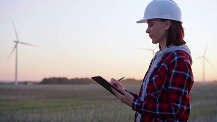 Woman engineer taking notes on a clipboard on a field with wind turbines, as the sun sets. Clean energy and engineering concept