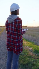 Woman engineer wearing a white protective helmet is taking notes with a clipboard in a field with wind turbines. Clean energy and engineering concept