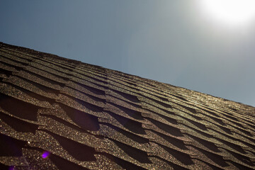 The morning sun casts a warm glow over textured roof shingles, highlighting their patterns and creating a shimmering effect under the clear sky.