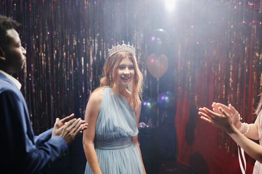 Excited woman in elegant blue gown and tiara standing in decorated party room smiling at camera under festive light