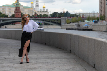 Full length portrait of beautiful blond long haired caucasian woman in eyeglasses, white shirt and black skirt standing on city street on river embankment in a cloudy day. Moscow Kremlin in background