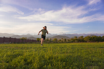 A bearded man with long red hair runs through a field, holding a phone in one hand. Wearing a green T-shirt, black shorts, and a cap, he is surrounded by trees and mountains under a wide sky.