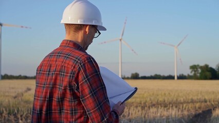 Man engineer, wearing a white protective helmet is taking notes with a clipboard in a field with wind turbines, as the sun sets. Clean energy and engineering concept