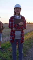 Woman engineer is standing straight and taking notes with a clipboard in a field with wind turbines, as the sun sets. Clean energy and engineering concept