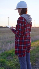 Woman engineer wearing a white protective helmet is taking notes with a clipboard in a field with wind turbines, as the sun sets. Clean energy and engineering concept