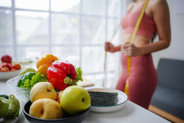 Woman is measuring her waistline in a bright room, with a selection of fresh fruits and vegetables...