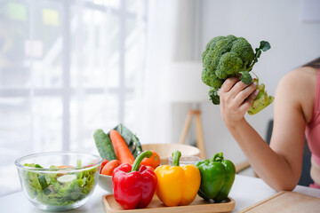 Young woman is holding a head of broccoli while preparing a healthy meal with fresh vegetables in her kitchen