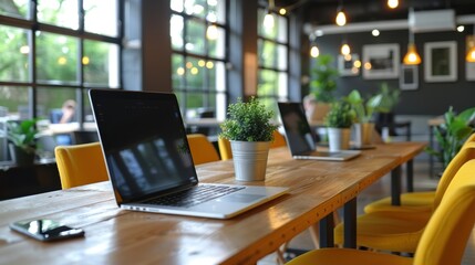 Fototapeta premium A modern, brightly lit co-working space with large windows, yellow chairs, and a wooden table adorned with laptops and small potted plants, creating an inviting work environment