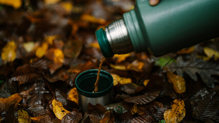 Close up of coffee pouring from green thermos into cup. Photo of metal stainless thermos lying on leaf in forest.