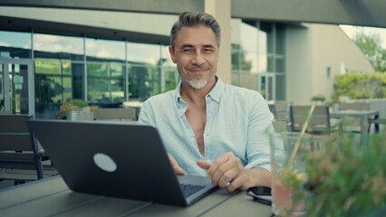 Smiling mature freelancer man working on laptop enjoying fresh air and freedom at restaurant terrace. Portrait of happy mid adult businessman. Male entrepreneur sitting at outdoor cafe.