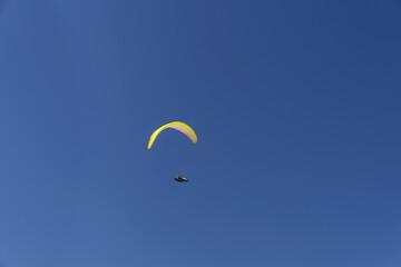 Paraglider soaring high in the sky with a yellow canopy, under a bright sun and scattered clouds.