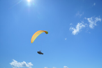 Paraglider soaring high in the sky with a yellow canopy, under a bright sun and scattered clouds.