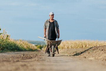 Senior Worker Walking with Wheelbarrow on Dirt Path Near Fields