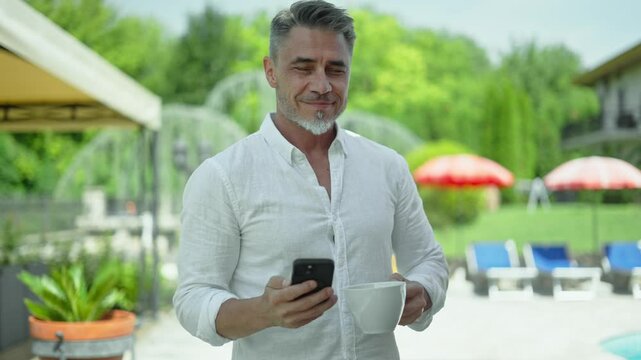 Older man drinking is morning coffee, checking phone outdoors. Handsome mid adult male with grey hair and white beard enjoying summer vacation at luxurious resort garden.