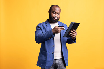 Irked man upset after lacking necessary money while trying to buy stuff online. Grumpy person looking at debit card after getting transaction rejected on tablet, isolated over studio background