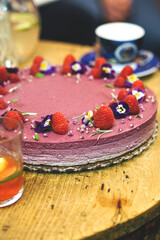 Close-up of a decorated berry cheesecake topped with fresh raspberries and edible flowers on a rustic wooden table. A cup and glass with a beverage are in the blurred background.