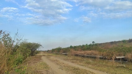 Obraz premium A dirt road meanders through a dry, rural landscape under a partly cloudy sky. The scene captures the quiet solitude of the countryside