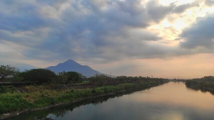 The big river surrounded with tree and bush then mountain as background during cloudy sunset