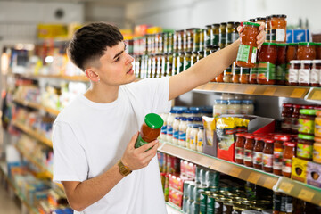 young man meticulously chooses tomato sauce in supermarket.