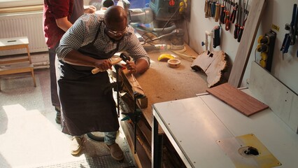 Sculptor shaping raw timber using chisel and hammer in carpentry shop with coworker cleaning workbench in background. BIPOC man making wood sculptures, engraving plank with tools, camera B