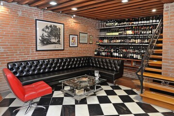 A stylish lounge area featuring a black leather couch and a wall of wines, set in a warm brick environment. Generative AI