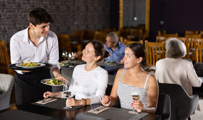 Skilled young waiter serving fresh vegetable salads to two women having friendly meeting in cozy...