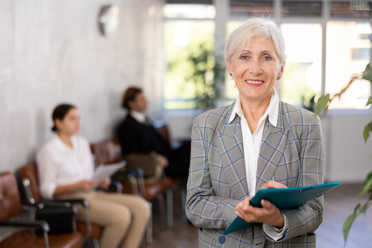 Friendly secretary senior woman stands at entrance to office reception with list of guests and expect invited visitors. Mature female office hostess willing to help