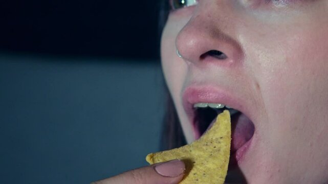 Macro view of woman opening mouth and biting nachos while watching interesting film on TV. Closeup view of satisfied female face eating crisps and enjoying exciting television program in the evening
